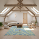 A styled living room in a bright attic with exposed wooden beams, featuring a blue and green patterned area rug, a cream sofa, a wooden coffee table, a woven chair, and a small white sheep figurine.