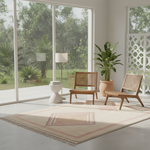 A styled interior scene with two wooden lounge chairs, a floor lamp, a potted plant, and a large patterned area rug in a bright room with tall windows overlooking greenery outdoors.