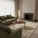 A modern living room scene featuring a sage green sofa, a matching armchair, a round wooden coffee table, and a large cream-colored area rug with a light green geometric pattern. The room is styled with neutral tones, a tiled fireplace, and soft natural light from a window with sheer curtains.
