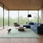 A modern living room with a large blue sectional sofa, a matching armchair, a low wooden coffee table, a light blue rug, and a black floor lamp, set against large windows overlooking a lush green outdoor landscape.