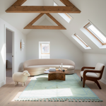 A styled interior room with a curved cream sofa, a wooden armchair, a light blue rug, and a small sheep-shaped stool, set in a bright attic space with exposed wooden beams and skylights.
