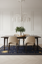 A styled dining room scene featuring a dark wood rectangular table, four upholstered dining chairs in a light beige fabric, a navy blue area rug with gold celestial patterns, and a white multi-arm chandelier hanging above.