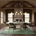 A styled dining room interior with a round white dining table, floral green upholstered chairs, a green patterned area rug, and a large black chandelier in a rustic wood-paneled room with a stone fireplace.
