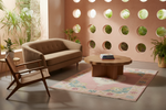 A styled interior scene with a beige tufted sofa, a wooden armchair, a round wooden coffee table, and a pink patterned area rug in a warm, modern room with a terracotta wall featuring circular cutouts.