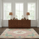 A pink area rug with a floral medallion and multicolor floral border is displayed on a dark wood floor in front of a brown wooden credenza, with two matching table lamps and decorative vases on top, set against a white paneled wall with three windows.