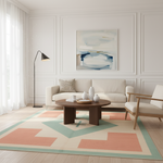 A styled living room interior featuring a large geometric area rug in beige, teal, and coral, a cream sofa, a round wooden coffee table, a black floor lamp, and a white armchair. A framed abstract painting hangs on the white paneled wall above the sofa.