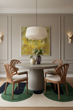 A styled dining room scene featuring a round gray concrete dining table with four wooden chairs with cream cushions, set on a green and navy patterned rug. A large pleated white pendant light hangs above, and a yellow abstract painting is mounted on the gray paneled wall behind.