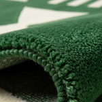 Close-up of a dark green textured rug with a raised loop pile border, folded over to show the underside and a portion of a white and green patterned surface underneath.