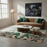 A styled living room featuring a large abstract art rug with green, pink, and cream wavy patterns. The rug is styled in a modern living room setting with a brown velvet sofa, green and patterned throw pillows, a white marble coffee table, a sculptural floor lamp, and a small cloud-shaped ottoman.