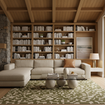 A modern living room featuring a large beige sectional sofa, a glass-top coffee table with round stone-like bases, a green and beige patterned rug, and a built-in wooden bookshelf filled with books behind the sofa.