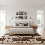 A modern bedroom interior with a bed centered against a white wall, framed black-and-white wall art above it, two wooden nightstands, black lamps, and a large geometric area rug in brown and cream. An open doorway on the right reveals another room with a chair and table.