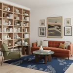 A styled living room interior featuring a terracotta orange sofa, a green armchair, a round marble coffee table, a blue patterned rug, and a large wooden bookshelf filled with books and decorative objects. Wall art hangs above the sofa.