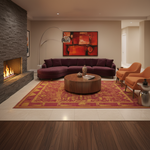 A modern living room featuring a plush purple sectional sofa, a round wooden coffee table, two orange armchairs, a large red and gold patterned area rug, and a stone fireplace on the left.