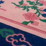 A close-up of a textured rug showing its detailed pile and pattern. The design features a light pink field with green and pink floral motifs, a pink border stripe, and a dark navy blue section at the bottom with pink cloud-like patterns.