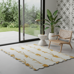 A rectangular area rug with a scalloped edge, featuring a mustard yellow floral pattern on a white background, styled in a modern room with a wooden chair, potted plant, and large glass doors.