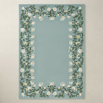 A rectangular light blue rug with a decorative border of white flowers and green leaves, shown flat against a neutral beige background.