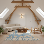 A styled attic living room with a curved cream sofa, a wooden lounge chair, a blue and cream patterned area rug, and exposed wooden beams on a white wall.