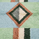 A close-up of a textured, shaggy blanket or rug featuring a geometric diamond pattern in light blue, mint green, dusty terracotta, dark green, and cream.