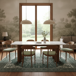A styled dining room scene featuring a rectangular white marble-top dining table with four wooden chairs that have light green cushions, set on a large green patterned area rug. The room has a warm wood-trimmed window, two dome pendant lights, and a sofa in the background.