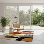 A modern abstract area rug with geometric shapes in red, yellow, black, white, and beige, styled in a bright, contemporary room with a wooden armchair, a potted plant, and a view of a lush green garden outside.