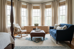 Living room with blue sofa, square blue rug, wooden armchair, and ottoman in front of large windows.