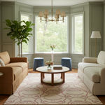A beautifully styled living room featuring a large beige and cream patterned area rug, a round wooden coffee table, two upholstered sofas, a bay window with lush green foliage outside, and a modern brass chandelier.