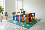 Colorful children's table and chairs set in a room with  green rug, large windows and plants.