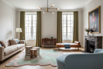 A beautifully styled living room featuring a beige sofa, a light blue armchair, a brown leather chaise lounge, a green patterned rug, olive green curtains, and a dark marble fireplace with a large portrait painting.