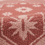 Close-up of a textured rug with a chunky, looped pile. The design features a geometric pattern in dusty terracotta red and pale blush pink, with a blurred background showing the same pattern.