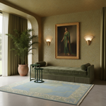A styled interior scene with a large framed portrait of a woman in a green dress hanging on a beige wall above a green daybed. The room includes olive drapes, a potted palm, two lit wall sconces, a blue patterned rug, and small green side tables.