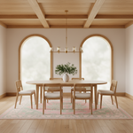 A minimalist dining room scene featuring a light wood oval dining table with six matching wooden chairs with white cushions, set on a pink and green patterned rug under a brass six-light chandelier. The room has a warm beige wall, arched windows with soft white light, and a light wood ceiling with exposed beams.