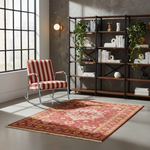 A red and beige patterned area rug with fringe is styled in a modern room with a red and white striped rocking chair, a large black-framed window, and a wooden bookshelf.