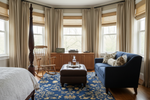 Living room with blue sofa, ottoman, blue floral traditional rug, and rocking chair near large windows with curtains.