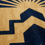 Close-up of a textured rug with a geometric pattern in tan and navy blue, featuring a sunburst and stepped house-like shapes.