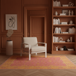A styled interior scene with a cream upholstered armchair centered on a pink and yellow patterned rug in a warm wood-paneled room. A bookshelf, framed wall art, and decorative objects are visible in the background.