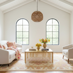A bright, modern living room featuring a light wood slatted coffee table in the center, surrounded by a beige sofa and armchair on a patterned rug. Two tall arched windows with black frames look out to green foliage, and a woven pendant light hangs from the high white ceiling.