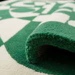 A close-up of a plush green rug with a rolled corner, showing its thick pile and texture, set against a matching green and off-white patterned background.