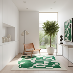 A modern living room corner featuring a prominent green and white abstract geometric area rug on a light wood floor. A mid-century modern wooden armchair sits on the rug next to a black floor lamp, while a large potted tree stands near a tall window.