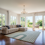 A modern living room featuring a beige tufted sectional sofa with blue and gray accent pillows, a striped area rug with a green arch motif, a crystal chandelier, and a hanging wicker chair overlooking a green garden.