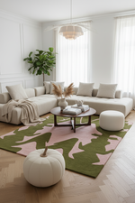 A styled living room featuring a large sectional sofa, a round wooden coffee table, a white pouf, and a green and pink camouflage patterned area rug on a light wood herringbone floor. A white decorative pumpkin sits in the foreground, and a potted green plant is in the background near white paneled walls and sheer curtains.