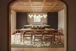 A styled interior dining room viewed through an arched doorway, featuring a wooden dining table and chairs on a green and white patterned rug, with a modern kitchen in the background.