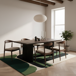 A modern dining room scene with a dark wood rectangular table and four matching chairs with light beige upholstered seats, arranged on a green, black, and cream abstract area rug. The room has light beige walls, a large window, a white pendant light, and a potted plant.