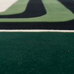 Close-up of a textured rug showing a dark forest green section in sharp focus, with a white dividing line and a blurred background of green and black curved stripes.