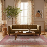 A styled living room scene featuring a tufted olive green sofa as the main product, placed on a patterned rug with a wooden coffee table, side table, floor lamp, curtains, and a potted tree in a bright interior.