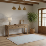 A styled entryway scene with a light wood console table, three brass pendant lights, a potted olive tree in a woven basket, and a pale blue rug with a pink geometric border on a light wood floor.