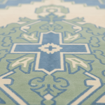 A close-up of a patterned rug featuring a geometric cross design in shades of blue, sage green, and cream.