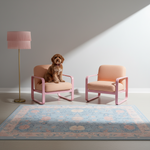 A small brown dog sits on one of two pink armchairs in a styled room with a patterned rug, a floor lamp, and strong diagonal sunlight on a light gray wall.
