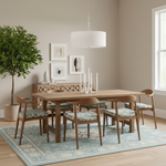 A dining room scene with a light wood rectangular table and six matching wooden chairs with blue floral upholstered seats, arranged on a pale blue patterned rug. A white drum pendant light hangs above, and the room has beige walls, framed abstract wall art, a sideboard, and a tall potted tree.