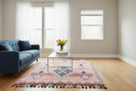 A pink and multicolor patterned area rug with tassels is styled in a bright, modern living room with a blue sofa, a glass-top coffee table, and light wood floors.