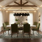 A staged dining room interior with a round white dining table, six olive green upholstered chairs, a large black circular pendant chandelier, a patterned area rug, and warm cream walls and curtains.